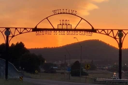 The Williams 1881 Gateway to the Grand Canyon arch in silhouette against an orange sunset sky, with the hills behind it